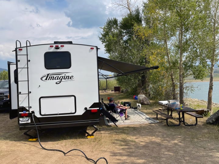 Relaxing at a scenic lake campsite with an RV rental, complete with an awning and picnic table.. Grand Design Imagine 2022