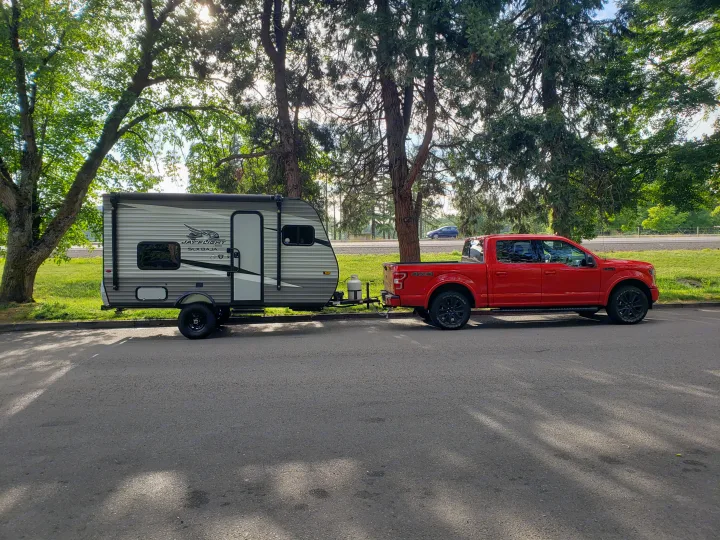 Red pickup truck towing a Jay Flight travel trailer under trees. Perfect setup for an RV rental camping trip!. Jayco Jay Flight SLX Baja Edition 2021