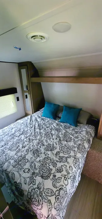 RV rental bedroom interior featuring a comfortable bed with a patterned comforter, blue pillows, and a window.. Keystone Hideout 2021