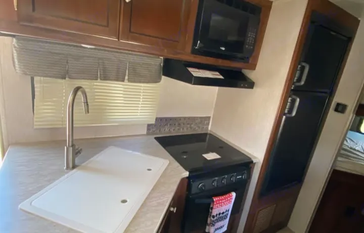 This RV rental kitchen features wood cabinets, a sink with a cover, a stove, microwave, and a black two-door refrigerator.. Heartland Other 2018