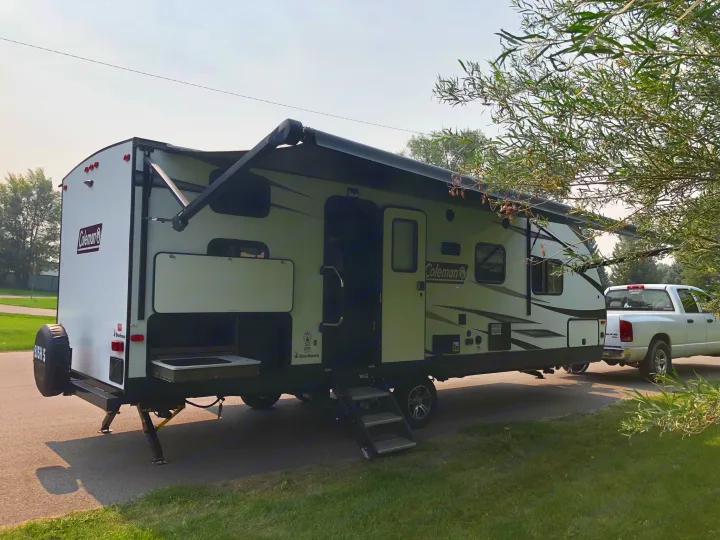 White & grey Coleman RV rental set up with an extended awning and outdoor kitchen, hitched to a white pickup truck on asphalt.. Coleman Other 2021