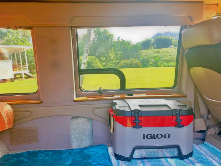 Inside an RV rental, a comfortable sleeping area with a blue blanket and a large Igloo cooler are visible, with lush green landscapes outside the windows.. Dodge Ram 1500 1999