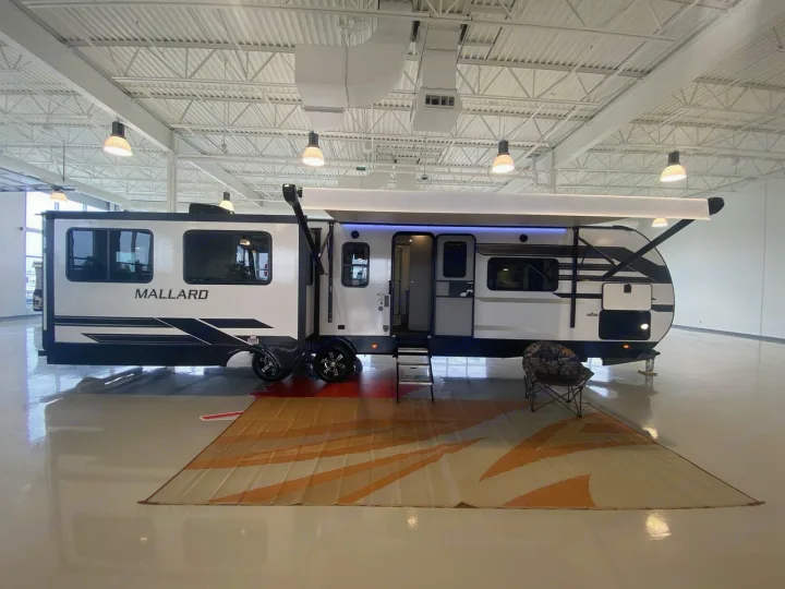 A sleek white and black Mallard RV travel trailer, ideal for rv rental, is showcased indoors with its awning and slide-out extended.. Heartland RVs Mallard 2021