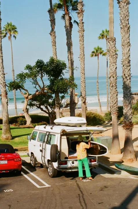 A white RV rental van with surfboards is parked on a sunny road near palm trees and the ocean. A person handles gear.. Volkswagen Westfalia 1986