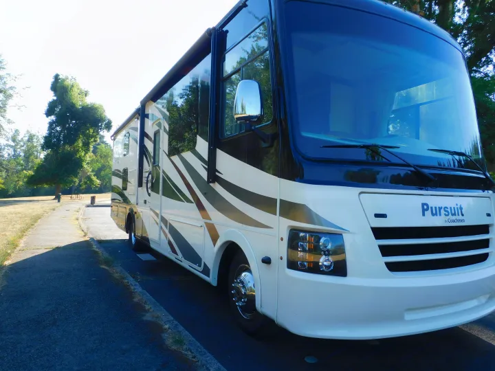 A white and black Pursuit by Coachmen RV rental is parked on asphalt, with its awning extended, ready for outdoor adventures.. Coachmen Pursuit 2017