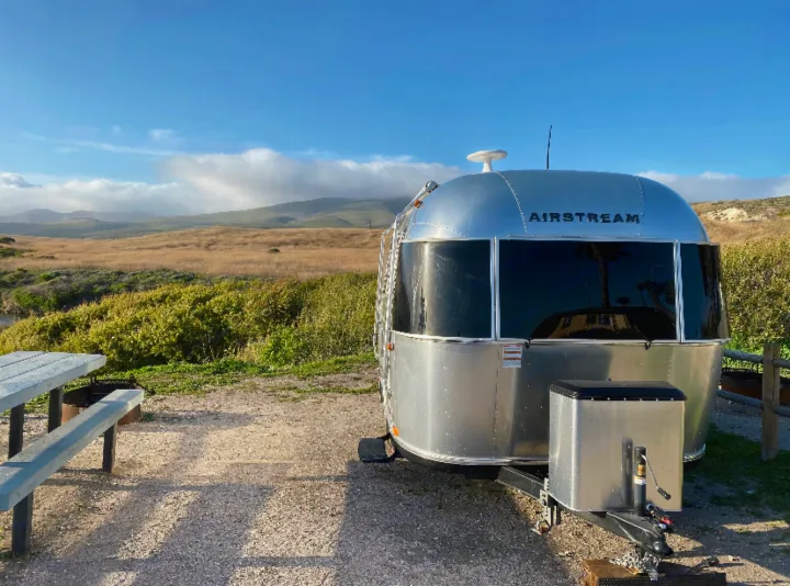 Airstream RV rental at a scenic campsite with a picnic table, featuring a natural landscape and distant cloudy mountains.. Airstream Bambi 2020