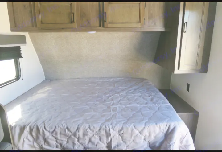 Inside an RV rental, a well-appointed bedroom features a large bed with grey quilt, overhead cabinets, and a side window.. Highland Ridge RV Open Range 2021