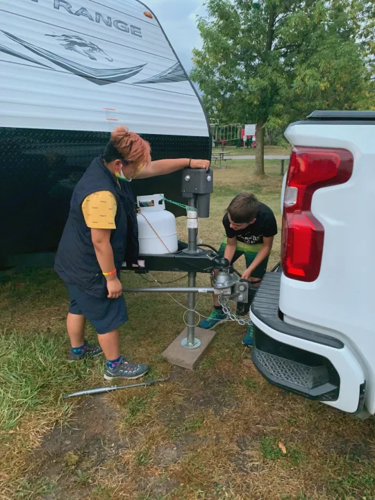 Two people preparing an RV rental: Hitching a white travel trailer to a pickup truck at a campground.. Highland Ridge RV Open Range 2021