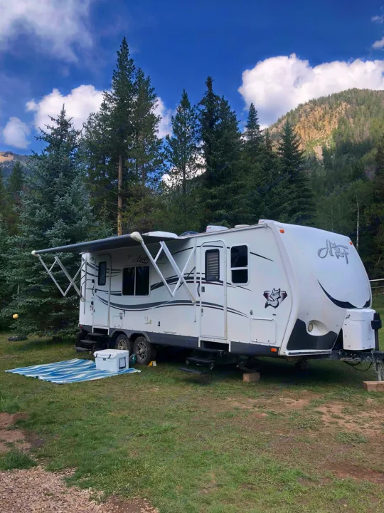 Serene RV rental campsite scene: white travel trailer, extended awning, cooler, and mat, surrounded by trees & mountains.. Northwood Mfg Arctic Fox 2013