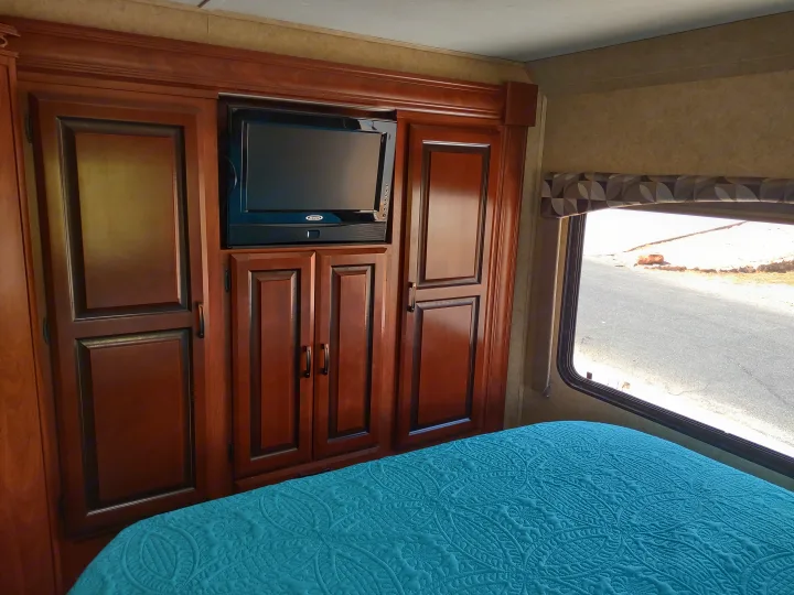 RV rental bedroom: rich wood cabinets, TV, vibrant teal quilted bed. Window shows road view outside.. Forest River Sunseeker 2012