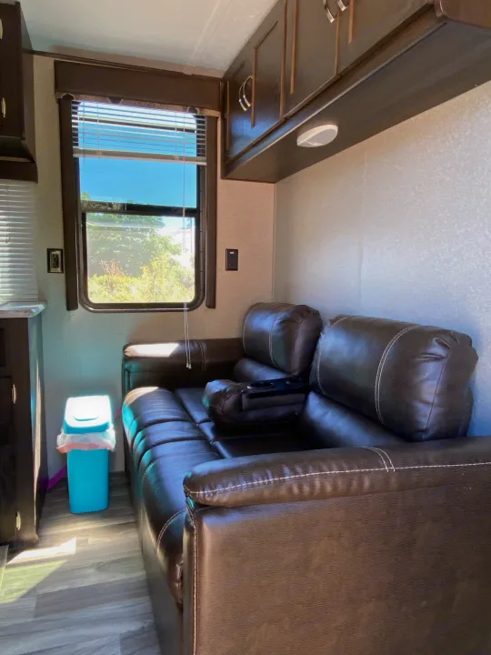 Cozy RV rental interior with a brown sofa, sunny window, overhead cabinets, and wood-look floor, perfect for relaxation.. Keystone Springdale 2018