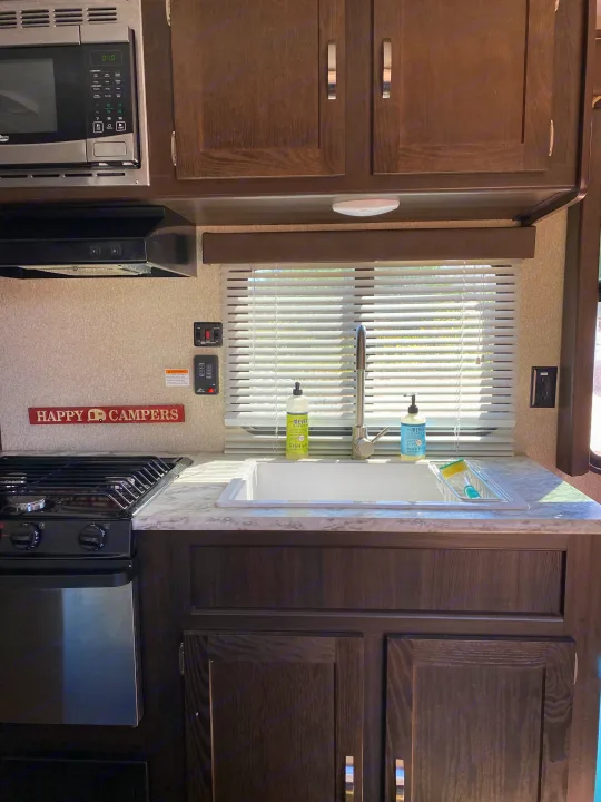 This RV rental kitchen features a microwave, stove, sink with soap, and dark wood cabinets, perfect for happy campers.. Keystone Springdale 2018