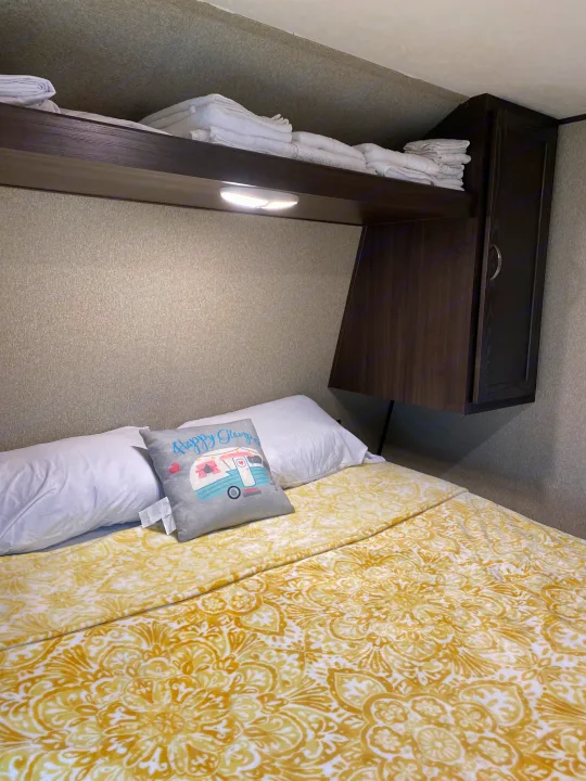 Cozy RV rental bedroom with yellow patterned bedding, white pillows, and overhead shelving for storage.. Keystone Springdale 2018