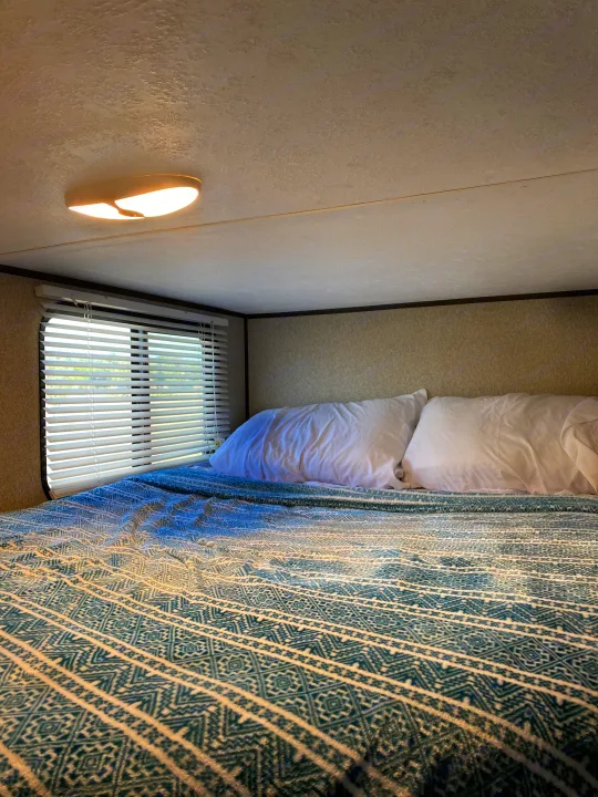 Cozy RV rental bedroom featuring a patterned blanket, white pillows, and a window with blinds, illuminated by a ceiling light.. Keystone Springdale 2018