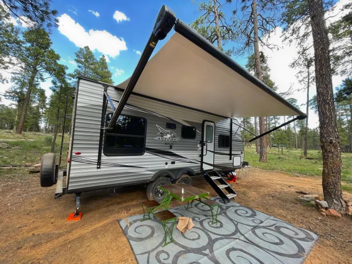 Enjoy nature with this RV rental travel trailer set up in a pine forest campsite, featuring an awning & patio rug.. Jayco Baja 2021