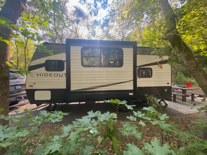 A "Hideout" travel trailer, a common RV rental, parked in a sunny, wooded campsite with a picnic table.. Keystone RV Hideout 2021