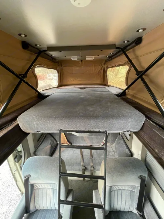 Upper sleeping berth of an RV rental camper van, accessed by a ladder. Features pop-top canvas windows and lower cabin seats.. Sportsmobile Custom Design 2013