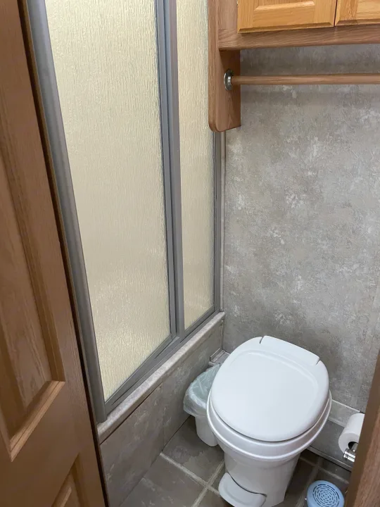 RV rental bathroom features a white toilet, compact shower with textured glass doors, and wooden overhead storage.. Concord Coachmen 2004