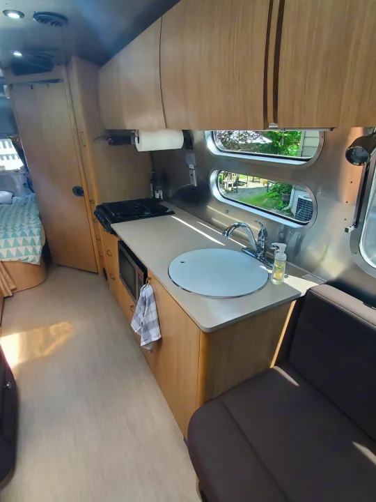 Bright RV rental interior showing a compact kitchen with sink, stovetop, and microwave, plus seating and a bed.. Airstream Flying Cloud 2018
