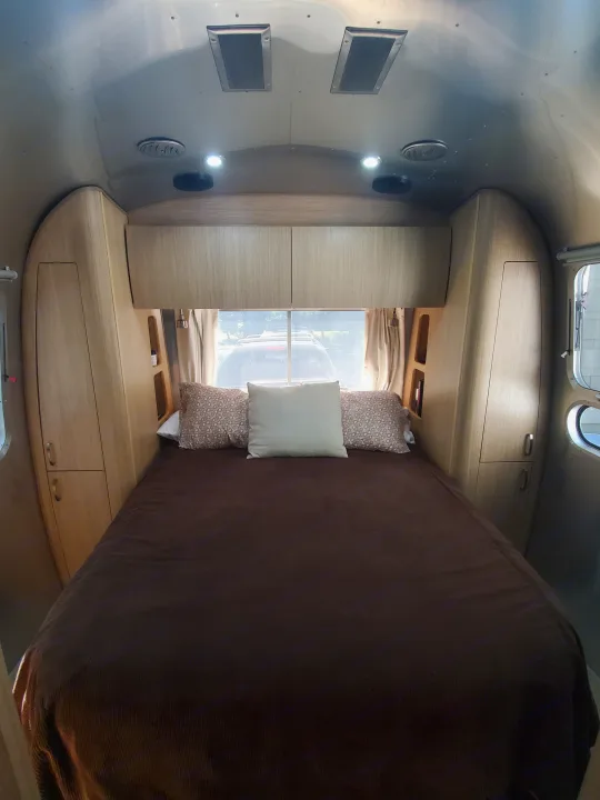 Cozy RV rental bedroom with a comfy bed, wood cabinets, and large rear window offering a view outside. Ideal for travel.. Airstream Flying Cloud 2018