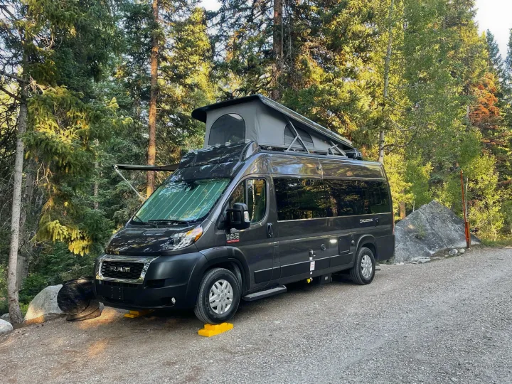 Enjoy nature with this dark grey rv rental camper van, parked in a sunny forest with pop-top tent extended for camping.. Thor Sequence 2022