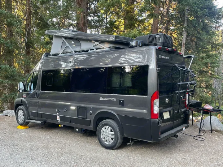 A gray pop-top camper van (RV rental) parked on gravel in a forest, with a grill set up for outdoor cooking. Ideal for camping travel.. Thor Sequence 2022