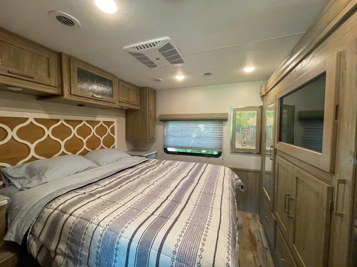 Cozy RV bedroom featuring a patterned comforter, overhead cabinets, and built-in TV. Ideal for your next RV rental.. Coachmen Pursuit 2021