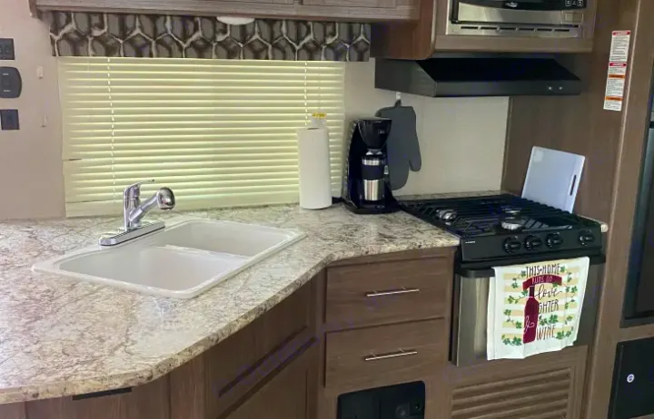 Inside an RV rental, a compact kitchen with sink, patterned countertop, stove, microwave, and coffee maker.. Keystone Hideout 2019