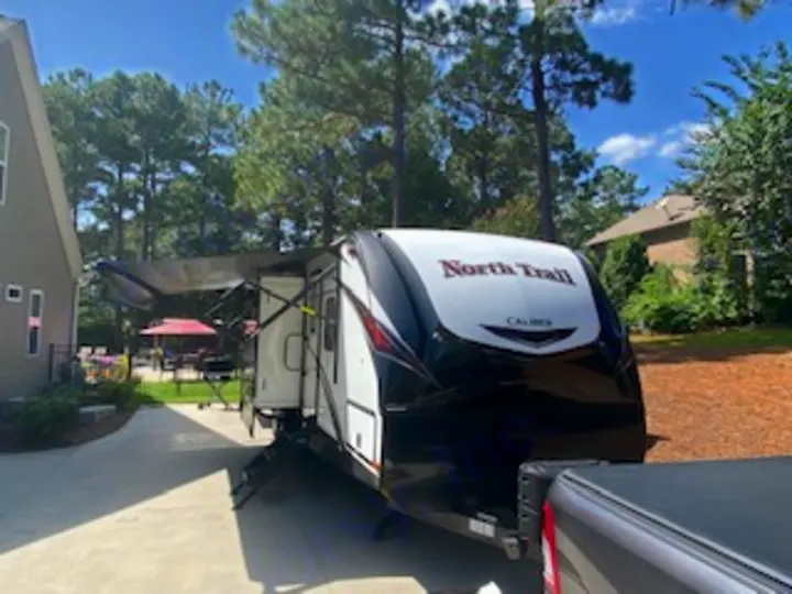 A white and black North Trail travel trailer RV, with awning extended, is parked in a driveway. Perfect for an RV rental.. Heartland RVs North Trail 2020