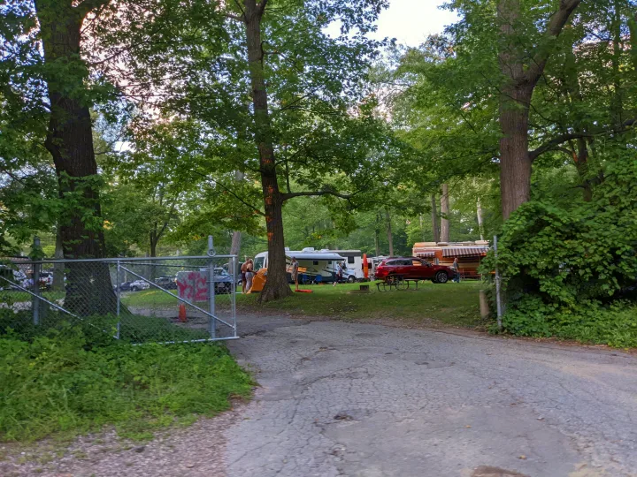RV rental site: RVs, a tent, people, and a red car seen through a fence at a shaded campsite.. Forest River Lexington 2008