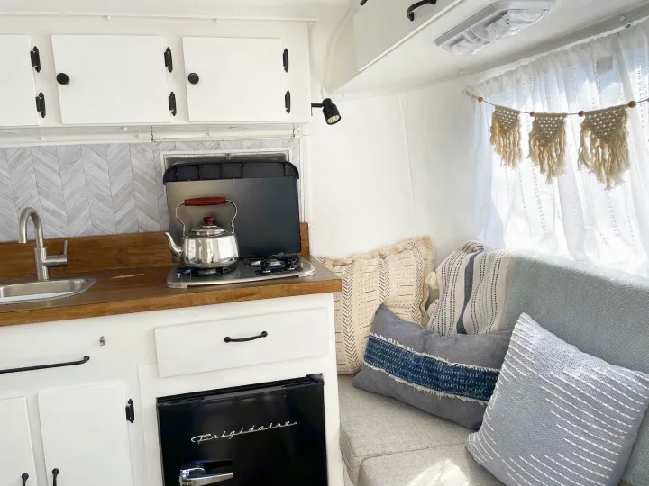 Bright RV rental kitchen with white cabinets, wood counter, stovetop, and cozy sofa with boho pillows. Clean, inviting space.. Scamp 13' 1979