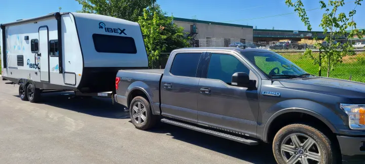 A gray Ford F-150 pickup truck tows a white Ibex travel trailer, ready for an RV rental camping trip.. Forest River All American Sport 2021