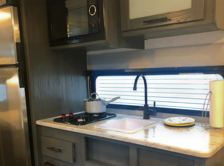 RV rental kitchen featuring a microwave, stove, sink, and stainless steel fridge. Dark wood interior and light counters.. Dutchman Aspen Trail 2022