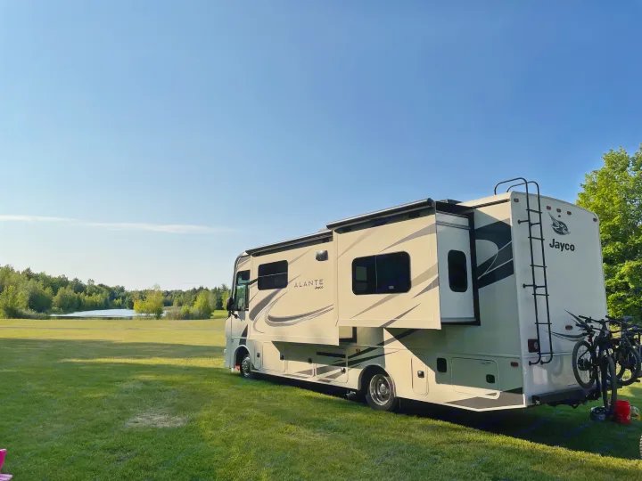 Enjoying an RV rental adventure. A large Class A RV is parked by a scenic lake, surrounded by green nature.. Jayco Other 2021