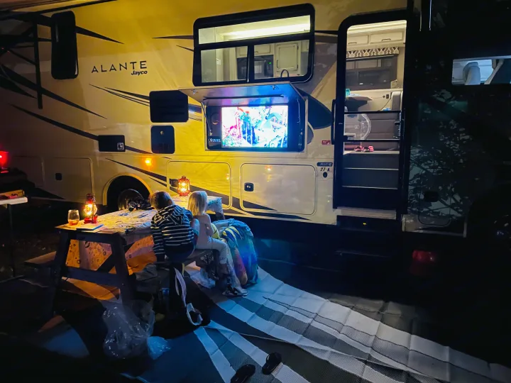 Nighttime family fun with an rv rental. Kids watch an outdoor TV by a lit picnic table and RV.. Jayco Other 2021