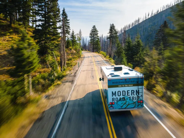 A white and blue Modern Road Trip RV rental drives on a scenic mountain highway, surrounded by green and burnt forests.. Winnebago Minnie Winnie 2021