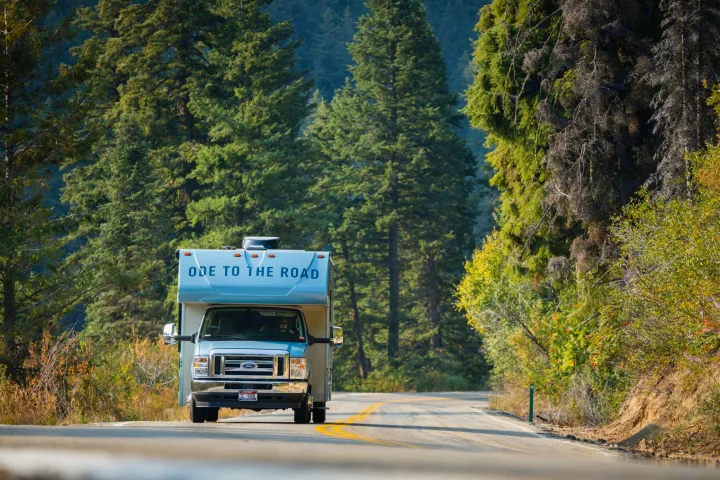 Light blue RV rental with 'ODE TO THE ROAD' drives on a scenic mountain road surrounded by lush green trees.. Winnebago Minnie Winnie 2021
