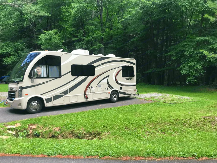 A beige RV rental, a Thor Vegas motorhome, parked on a pad in a lush green forest campground, with a person smiling inside.. Thor Vegas 2016