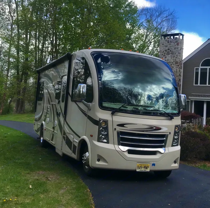 A large beige RV, with clouds reflecting on its windshield, is parked on a paved driveway. Consider it for an RV rental.. Thor Vegas 2016