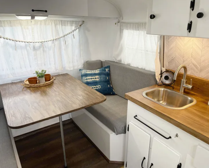 Bright RV rental interior: wooden table, gray bench, and mini kitchen with sink. White curtains & light wood create a cozy, clean space.. Scamp 13' 1979