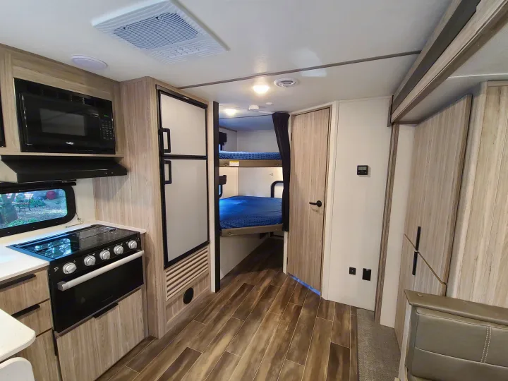 Modern rv rental interior: kitchen with microwave, stove, fridge; cozy bunk beds visible in the back, light wood cabinetry.. Keystone Passport 2021