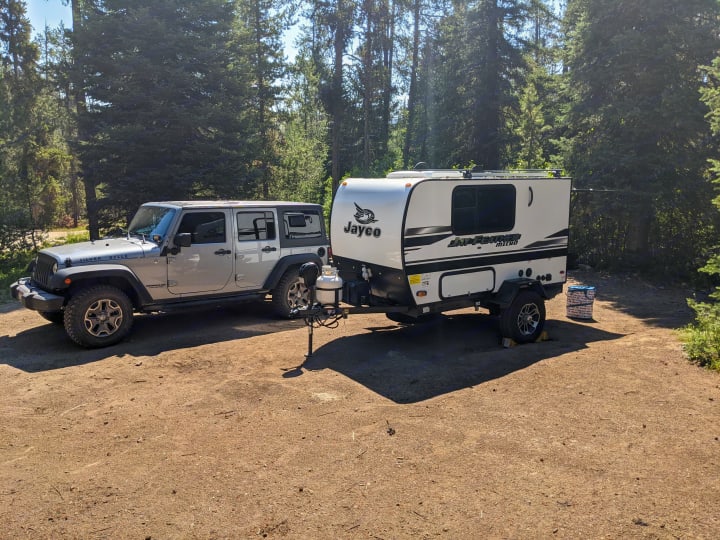 Jeep pulling a Jayco Jay Feather Micro RV camper at a sunny forest campsite. Great rv rental for adventure.. Jayco Jay Feather Micro 12SRK 2021