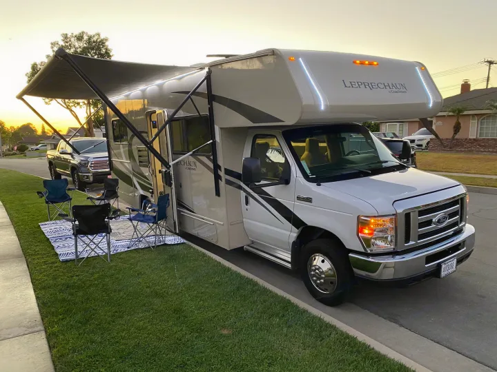 White RV rental (Coachmen Leprechaun) parked at sunset. Awning extended with outdoor chairs & mat on the grass.. Coachmen Leprechaun 2021
