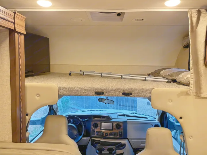 Inside an RV rental: the driver's cockpit, overhead bunk with ladder, TV, and ceiling lights/vents.. Thor Motor Coach Chateau 2017