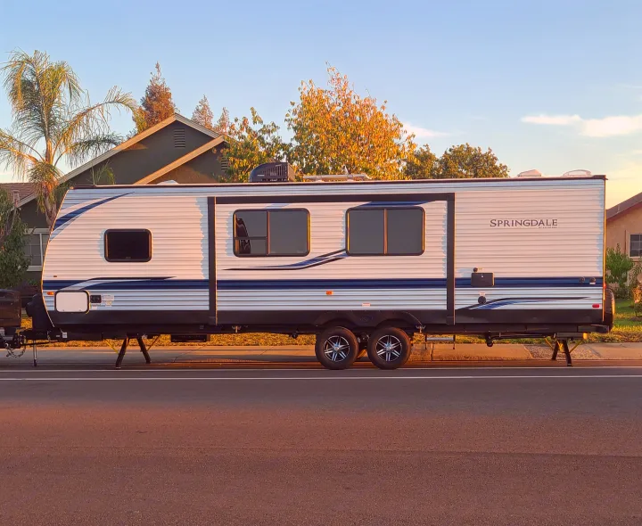 A large white Springdale travel trailer, an ideal rv rental, is parked roadside. It has blue stripes, windows, and dual axles.. Keystone Springdale 2022