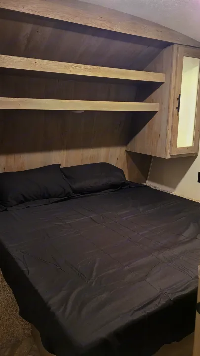 Cozy RV rental bedroom featuring a bed with dark sheets, built-in wooden shelves, and a mirrored cabinet.. Keystone Springdale 2022