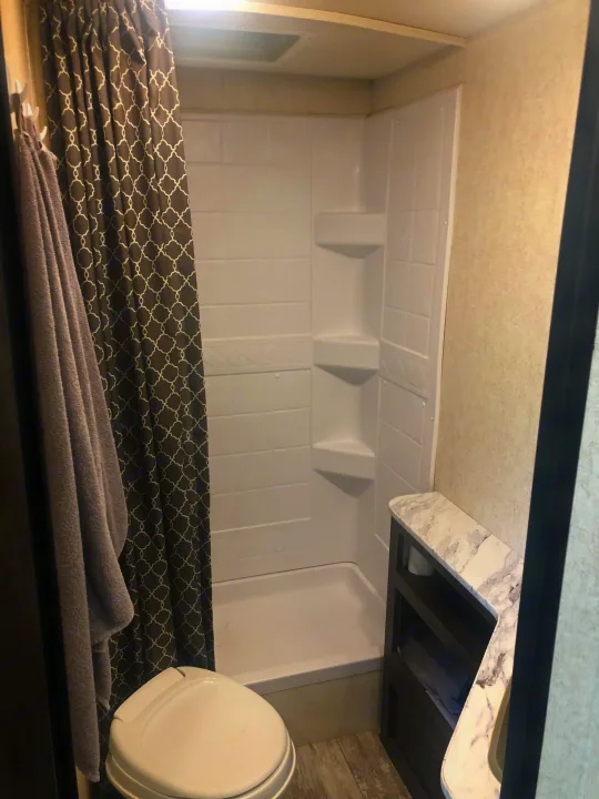 RV rental's bathroom features: white shower with built-in shelves, toilet, gray patterned curtain, towel, & marble vanity.. Jayco Highland Ridge 2019