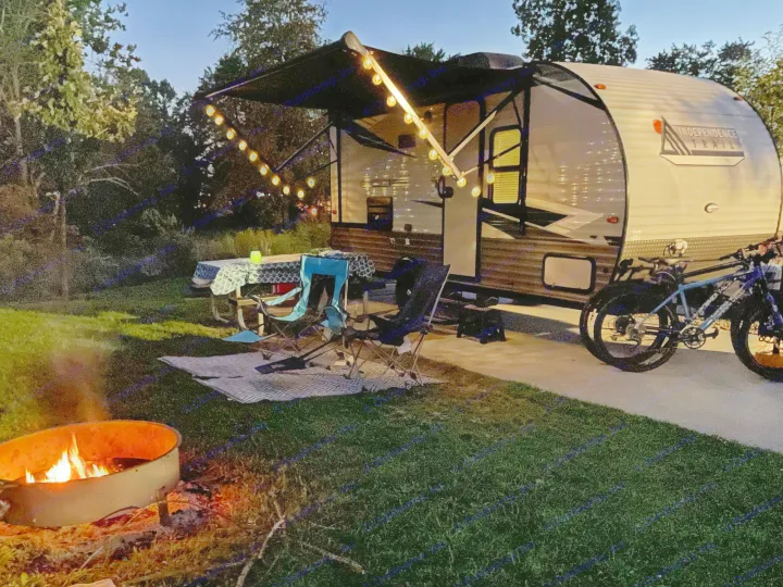 An RV rental campsite at dusk, featuring a cozy campfire, string lights, and bicycles next to the travel trailer.. Forest River Independence Trail 2021