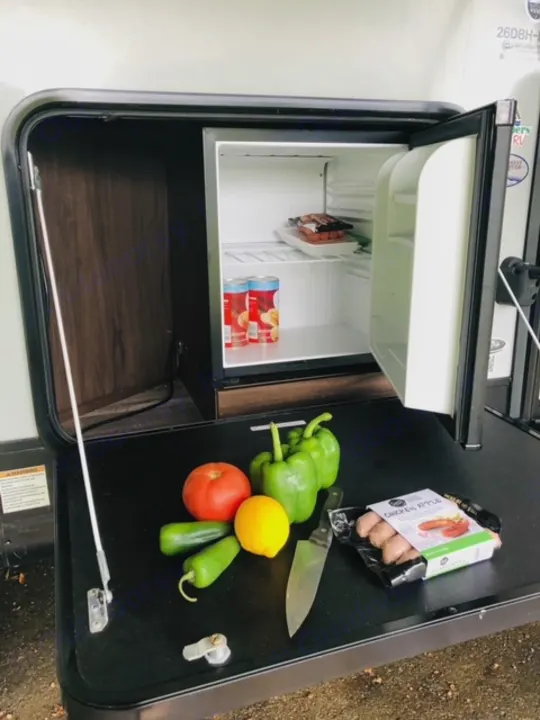 RV rental outdoor kitchen with open fridge (sausages, cans), counter with tomato, peppers, jalapeños, knife & chicken apple sausage.. Forest River Cherokee Alpha Wolf 2018