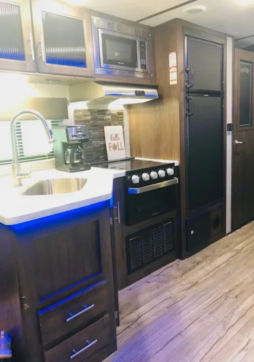 Inside an RV rental: a compact kitchen with modern sink, oven, microwave, fridge, and blue under-cabinet lighting.. Forest River Cherokee Alpha Wolf 2018